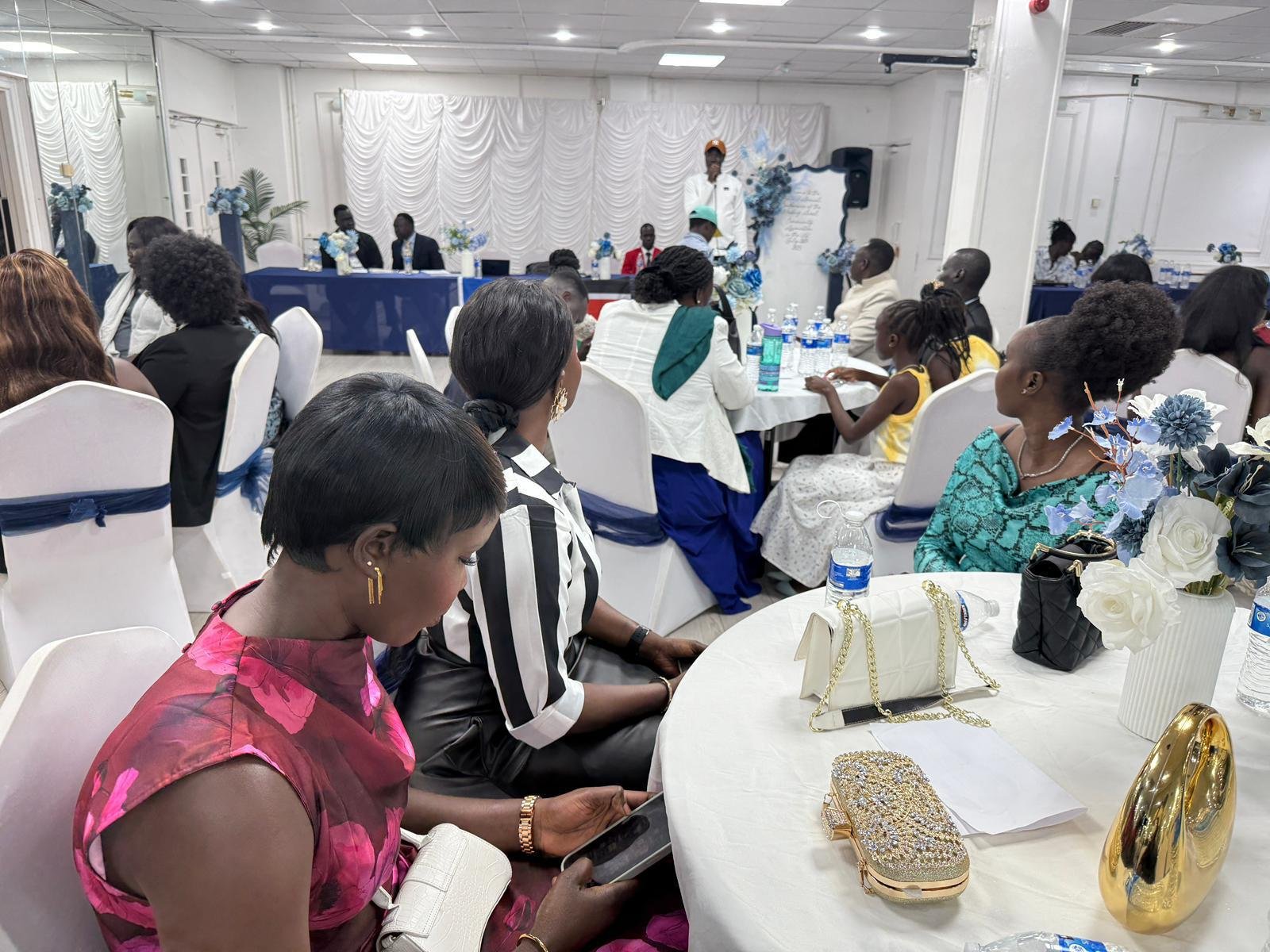 Image of community members at July 2025 gathering featuring a youth leader speaking at the front, with varied attendees seated at tables, including a woman, man, and child at two tables.